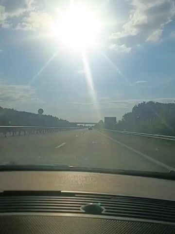 The video shows a view from inside a car driving on a highway. The sun is bright and high in the sky, casting a glare. There are other cars on the highway, including a dark-colored car in the left lane that is overtaken. The environment appears to be rural with trees and vegetation on the sides of the road. The road is paved and has multiple lanes. The video is likely recorded during the daytime.