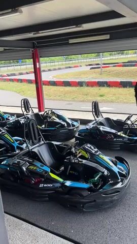 The video shows a go-kart track. In the first shot, several black and blue go-karts are parked under a covered area. The track is visible in the background. The camera moves to show a closer view of the karts, and then a person in a black long-sleeved shirt appears in the frame, standing on the track. One go-kart is parked on the track outside the covered area. The background shows the go-kart track and some green trees and hills in the distance.