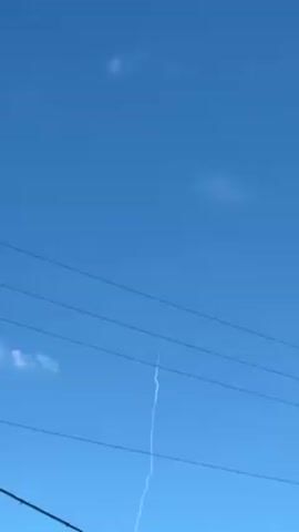 The video shows a bright blue sky with wispy clouds and a long, thin white contrail bisecting the frame vertically. The contrail appears to originate from a point high in the sky and descends towards the horizon. In the foreground, a series of dark power lines stretch across the bottom of the frame. The camera is positioned at a low angle, looking upward.