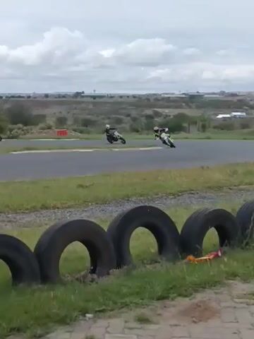 Two motorcyclists are racing on a paved track. The rider closer to the camera is on a black and white motorcycle and leans to the left as they round a curve. The other rider is on a red motorcycle. The track is surrounded by green grass and a row of black tires. The background shows a flat, grassy landscape under a cloudy sky.