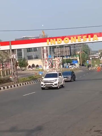 The video begins with a view of cars driving on a road. A large overhead sign with "BOGOR" and "INDONESIA" is visible. The sign is red and white, with the Indonesian flag colors. The scene then shifts to a different section of the road, showing a sign that reads "KABUPATEN". More cars are visible, and lush green trees line the roadside. The camera pans to show a tall, slender building with multiple windows, resembling a tower. In front of the tower, there is a sign for "PULLMAN" and "PULMAN HILLS". The environment appears to be tropical or subtropical, with palm trees and dense vegetation. The sky is clear and blue, suggesting daylight. The overall impression is of a scenic drive through an urban or semi-urban area with notable landmarks.