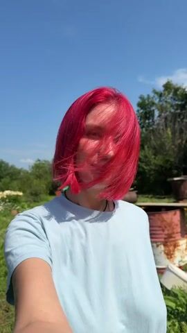 A person with short, bright pink hair is standing outdoors, possibly in a garden or yard. They are wearing a light blue t-shirt. The person is looking at the camera and smiling. The background is a bright blue sky with some greenery and trees. There are flowers and some objects that appear to be garden-related in the background. The lighting suggests it is a sunny day.