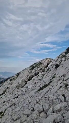 The video shows a panoramic view of a mountainous landscape. The foreground is dominated by rocky terrain, with patches of sparse green vegetation. In the mid-ground, rugged mountain ridges are visible, some covered in white rock and others with dark green forests. Sunlight casts shadows on the slopes, highlighting the texture of the rocks and foliage. In the distance, layers of hazy blue mountains recede towards the horizon. The sky is partly cloudy, with streaks of white and gray clouds against a pale blue backdrop. A faint rainbow is visible in the distance, arcing across the sky over the mountains. The camera is positioned at a high vantage point, looking down and across the vast expanse of the mountain range. The overall impression is one of rugged natural beauty and serene wilderness.