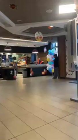 The interior of a McDonald's restaurant is visible in the video. The restaurant appears to be mostly empty, with clean, tan tiled floors. There is a counter area where food is prepared and served, with employees visible behind it. A woman stands holding a large bunch of pastel-colored balloons near a wall, looking at her phone. The restaurant is decorated with Halloween-themed decorations, including hanging spiders and pumpkins. Overhead lights illuminate the scene, and there are digital menus on stands.