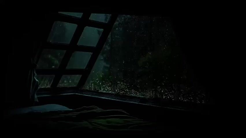 The video shows a view through a window at night. The window is divided into diamond shapes by a dark frame. Raindrops are visible on the glass, streaking down. Outside the window, trees and foliage are dimly visible, shrouded in darkness and rain. In the foreground, a bed with green blankets and white pillows is partially visible. The scene is dark and atmospheric, suggesting a cozy or perhaps isolated setting.