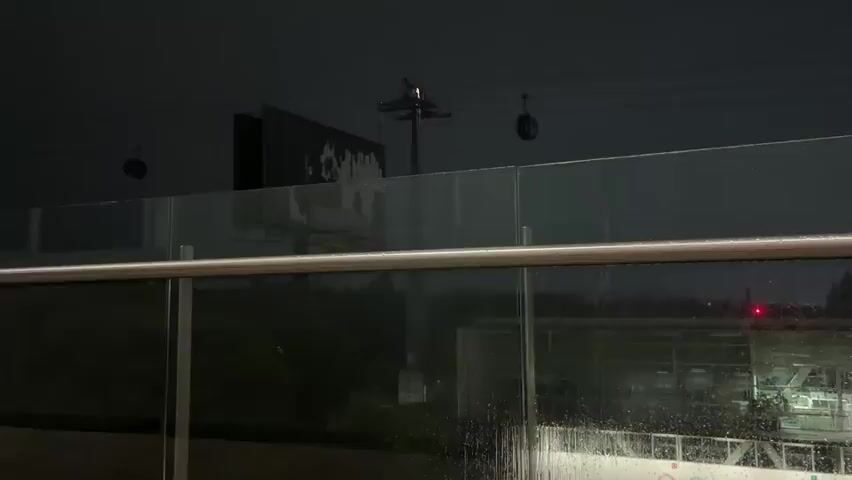 The video opens with a view of a dark, rainy night from an elevated outdoor position, likely a balcony or walkway. Heavy rain streaks are visible on a glass railing with a metal handrail. In the distance, two cable car gondolas are seen against the dark sky, along with a tall utility pole or tower and a large, dark rectangular billboard-like structure. A faint red light is visible in the background.

The camera then pans down to show an empty aluminum can lying on a concrete floor. Following this, there's a brief close-up of a person's face (likely the videographer), with dark, curly hair and eyes, illuminated by a reddish light. In the background, modern building architecture with recessed overhead lighting is visible. The video concludes by returning to the initial dark, rainy outdoor view through the glass, with raindrops and blurred distant lights dominating the frame, emphasizing the inclement weather.

The scene depicts a rainy night in an urban environment, possibly in Mexico City, given the presence of cable cars, which are a feature of the city's transport system. No discernible text is visible on any objects.
