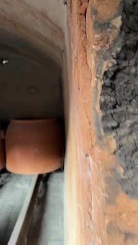 The camera pans across a small, enclosed space with a low, arched ceiling, possibly an oven or kiln. Inside, there are several large, reddish-brown ceramic pots stacked and arranged on a wooden track. The walls of the enclosure are made of brick or stone, and the ceiling appears to be made of a rough, textured material. There are a few small lights visible on the ceiling. The overall lighting is dim, and the space appears to be somewhat cramped. The objects are static, with no visible people or interactions taking place.