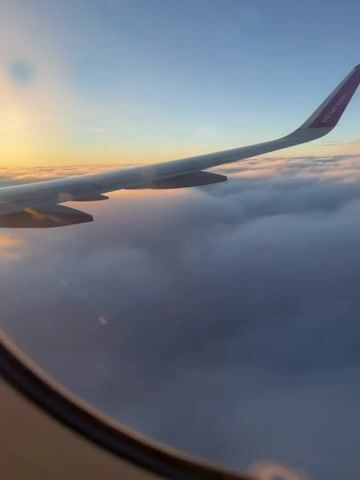 This is a first-person perspective video taken from an airplane window. The camera is angled towards the right, showing a portion of the airplane wing and the sky. The wing is a light gray with the airline's logo, which appears to be pink, visible on the wingtip. Below the wing, a thick layer of clouds stretches across the horizon. The sky is a gradient of blue and transitions to a warm orange and yellow hue on the left side, indicating either sunrise or sunset. The lighting suggests it is either early morning or late evening. The clouds are dense and appear fluffy, with some areas illuminated by the sun. The airplane window frame is visible in the foreground, creating a tunnel-like effect. The video is static, with no noticeable movement other than the subtle shifts in light and shadow on the clouds.