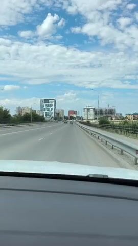 The video shows a first-person view from inside a car traveling on a highway. The highway has multiple lanes and appears to be elevated, with a guardrail on the right side. In the distance, there are several modern buildings, including skyscrapers under construction. The sky is blue with scattered white clouds. The car's dashboard is visible in the foreground. The traffic on the highway is light.