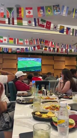 The video shows a group of people seated at tables in what appears to be a restaurant or sports bar. The room is decorated with numerous national flags hanging from the ceiling in garlands. A large television screen is mounted on the wall, displaying a soccer match. Several people are visible in the foreground, some looking at the screen, others at their phones or engaged in conversation. Food and drinks are present on the tables. The lighting suggests it is indoors, possibly during the day or evening.