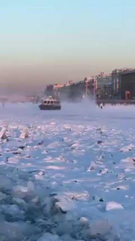 The video shows a hovercraft moving over a frozen river. The river is covered in snow and large chunks of ice. The hovercraft kicks up a cloud of snow and ice as it moves. In the background, there is a cityscape with buildings, and a few people can be seen walking on the frozen river. The sky is clear with a hint of color from the sunset or sunrise.