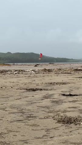 The video shows a beach with a red flag on a pole in the distance. The beach is covered in debris, including plastic bottles, wrappers, and other trash. The water in the background is murky and there are waves. The sky is overcast.