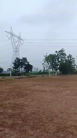 A video shows an outdoor scene with a large metal power line tower on the left side of the frame. The tower is tall and has a lattice structure. Power lines extend from the tower across the frame. In the middle ground, there are trees and a soccer goal. On the far left, a house with a red roof is visible. The ground in the foreground is brown and appears to be dirt or dry grass, with some scattered white debris. The sky is overcast and gray. The camera is static and facing the scene.