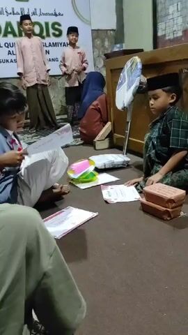 The video shows a group of children sitting on the floor in a room. There are two children standing in the background, looking at a banner. One of the children is holding a microphone. There is a fan in the background. There are papers and books on the floor. The children are wearing traditional clothing. The room appears to be a classroom or a place of worship. The overall atmosphere is calm and studious.