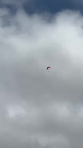 A single paraglider with a red canopy and a dark pilot is visible in the sky against a backdrop of overcast clouds. The paraglider is positioned in the upper center of the frame and appears to be in stable flight. The surrounding clouds are a mix of gray and white, with some darker areas suggesting thickness. The sky above the clouds is a deep blue. The video captures a static shot of this scene for approximately 4 seconds.