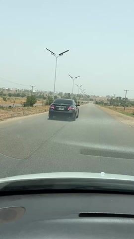 The video shows a view from the dashboard of a moving car, driving on a paved road lined with tall street lights. A black car is visible in front, moving in the same direction. The environment appears somewhat arid, with sparse vegetation along the roadside and buildings visible in the distance. The sky is overcast, and there are some utility poles and lines visible in the background. The car's dashboard is in the foreground, occupying the lower portion of the frame.