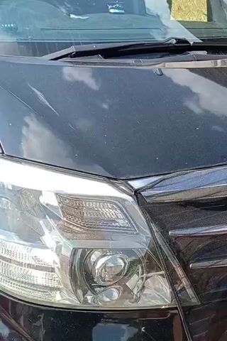 The video shows a close-up, side view of a black car parked outside. The camera is focused on the front passenger side of the vehicle, capturing the headlight, fender, and the front edge of the door. The car's surface appears to have dust and some water splashes. Reflections of the sky and clouds are visible on the car's hood. The background is blurred, but suggests an outdoor environment with some fencing and foliage. The video is very short and shows no people or any action.
