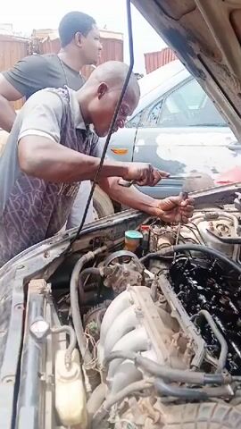 Three individuals are shown working on the engine of a car. The primary focus is on a man in the foreground, wearing a grey t-shirt with a dark pattern, who is using a tool to adjust something within the engine. He appears to be concentrated on his task. To his right, another man, wearing a red shirt, is also engaged in working on the engine, using a wrench. In the background, a third individual, wearing a dark t-shirt, is looking on. The car's hood is open, revealing the complex mechanical components of the engine, which appears somewhat dirty and greasy. The scene suggests a mechanic's workshop or an informal repair setting, given the clutter and the nature of the work being performed.