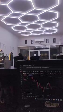 A short video clip shows the interior of a room with a unique hexagonal lighting fixture on the ceiling. In the foreground, a laptop screen displays a stock trading chart. The chart shows a downward trend in the stock price. In the background, several people are visible, some appearing to be engaged in conversations. The room appears to be a modern office or workspace, with clean white walls and large windows. The overall atmosphere is one of focused work or contemplation.