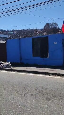 The video shows a street scene in what appears to be an urban area, possibly a neighborhood in a city or town. Along the side of the road, there is a large pile of trash consisting of bags, boxes, and assorted items. The trash is located on the sidewalk next to a blue wall and a closed metal door.

In the video, several individuals wearing what appears to be uniform or work attire, including white hard hats and tan suits, walk along the sidewalk. They each carry a blue bag or pack on their backs and are walking in the same direction. The group is walking in a coordinated manner.

The background of the scene includes buildings, fences, and a hillside with dense housing. In the distance, there is a restaurant sign visible. The weather appears to be sunny and clear, with a bright blue sky. Power lines are visible overhead.