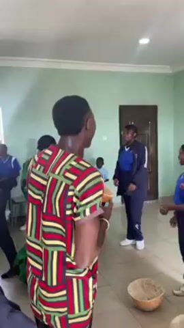The video shows a group of people in a room. One man in a colorful patterned shirt is singing loudly and gesturing with his hands. Several other people are standing around him, some clapping along. A woman in a blue tracksuit and glasses is dancing. A man is holding a percussion instrument and another man in a checkered shirt is clapping. The room has a light-colored wall and tiled floor. There is a dark door in the background. The overall scene appears festive and energetic.