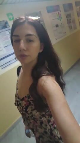 A young woman with dark hair is walking down a hallway. She is wearing a floral print dress and has sunglasses on top of her head. There are posters on the walls of the hallway.