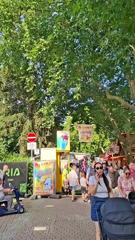 The video opens on a sunny day with a tree-lined path or sidewalk. A woman in a blue skirt and white top walks past what appears to be an outdoor event or fair. To her left, a man in a wheelchair is parked. Behind the wheelchair, a banner with a green arrow and running man symbol is visible, along with a black banner that says "KORA Kfz-Gutach ten." Further down the path, there are various stalls, possibly selling food or drinks, with colorful posters. People are milling around these stalls. The camera pans to the left, showing more trees and a blue sky with some clouds. A "no entry" sign with a bicycle symbol is mounted on a pole. Beyond this, a group of people are seated at picnic tables under a covered structure. The camera then pans slightly to the right, showing more buildings and trees. The video appears to capture a casual outdoor gathering.