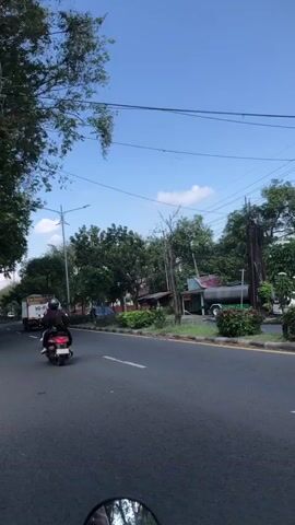 The video shows a view from the perspective of a motorcycle rider on a paved road. In the foreground, the reflection of the rider and the motorcycle handlebars are visible in a rearview mirror. The road ahead is asphalt, with a yellow painted line on the right side. A motorcyclist, wearing a dark helmet and jacket, is riding in front of the camera. To the left of the road, there are trees and a small truck. To the right, there are more trees, some bushes, and a small building with a pink roof. A large tank on a stand is also visible to the right, behind some foliage. The sky is blue with a few wispy clouds. There are utility poles and wires above the road. The overall environment appears to be an urban or suburban area on a clear day.