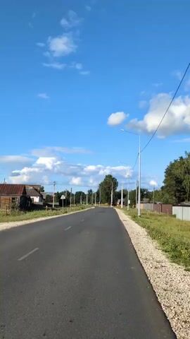 This video shows a straight, paved road in a rural area. The road is lined with streetlights on both sides. There are houses and trees along the sides of the road. The sky is blue with some white clouds. The video appears to be filmed from a moving vehicle, as the perspective of the road and scenery changes. There are no people or animals clearly visible in the video.