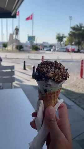 A person's hand holds an ice cream cone in a waffle cone. The ice cream has two visible layers: a green layer at the bottom and a white layer on top. The white layer is topped with chocolate and nuts. The ice cream cone is partially wrapped in white paper. In the background, there is a street with cars and pedestrians. There is also a statue and two Turkish flags visible. The setting appears to be outdoors, possibly a promenade or a public square, with a bright, sunny sky. The camera is focused on the ice cream cone.