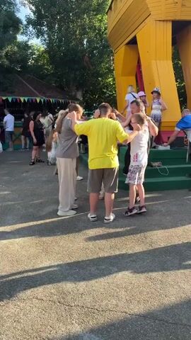 A group of five people are dancing in a circle outdoors on a paved surface. The person in the center is wearing a bright yellow t-shirt and gray shorts. To their left, a person in a gray t-shirt and light-colored pants is facing inward. To the yellow-shirted person's right, a person in a white t-shirt with a pattern and patterned shorts is dancing. Behind the person in the yellow shirt, a person wearing black is also dancing. To the yellow-shirted person's left, another person is partially visible, also dancing. In the background, several other people are standing and watching. There is a colorful structure with a roof and some stalls to the left, and a large yellow wooden structure resembling a castle or ship to the right. Trees and foliage are visible in the background. The sun is casting long shadows on the ground.