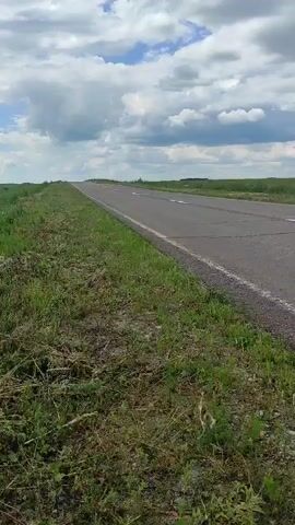 The video shows a landscape view of a grassy field with a road running alongside it. The sky is partly cloudy. There are no people or vehicles visible in the video. The camera appears to be moving, likely being held by someone walking or riding in a vehicle, as the perspective shifts slightly. The focus is on the natural environment, with the road acting as a boundary to the field. The grass is green and appears to be growing wild in some areas, with some dry vegetation also visible. A line of trees can be seen in the distance on the left side of the frame.