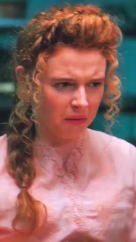 A close-up shot focuses on a woman with auburn curly hair styled in an updo with a braid. She is wearing a pink ruffled blouse and a dark skirt. Her facial expression shifts from concern to a slight smile. The background is blurred, suggesting an indoor setting.