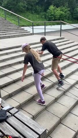 A group of children are engaged in a stair climbing exercise outdoors. They are tethered together by resistance bands, with some children pulling and others pushing. The children are of various ages and are dressed in casual clothing and athletic wear. The environment appears to be a public space with concrete stairs and a paved area at the top. There are trees and buildings visible in the background. The overall scene depicts a physically active group participating in a coordinated exercise.