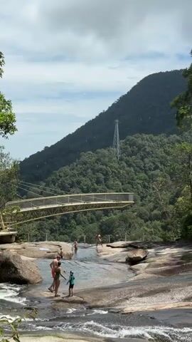 The video shows a natural landscape with a waterfall and a bridge. Several people are visible. In the foreground, a person is lying on a rock near the water. To the left, a group of people are standing in the shallow water of the waterfall. In the background, a suspension bridge crosses over the waterfall, and a tall metal tower is visible on a forested mountain. The sky is cloudy.