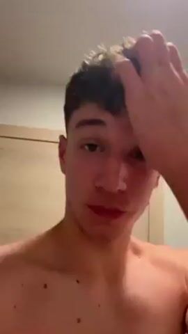 A shirtless young man with short curly brown hair is looking directly at the camera, running his hand through his hair. He is indoors, with a light brown door visible in the background. He appears to be in a bathroom or bedroom, with a white wall and a blue towel visible. He is mouthing words.