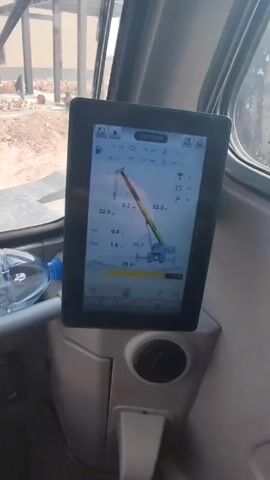From inside a crane, the video shows the crane lifting a metal beam to a steel structure under construction. The crane operator is using a monitor to view the load and crane status. The structure has multiple columns connected by beams, and some workers are visible on the construction site. The ground is dirt and rubble. A water bottle is visible in the crane cabin.