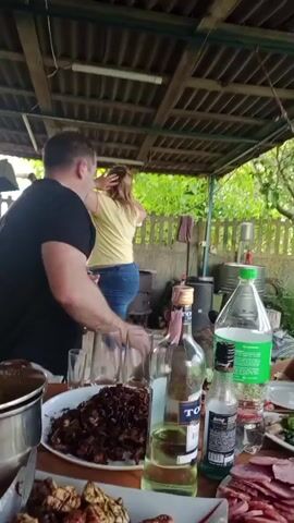 The video shows a man in a black t-shirt with a red and white logo on it, sitting at a table with food and drinks. He is holding a brown bottle in his left hand and a bottle opener in his right hand, appearing to open the bottle. In the background, a woman with blonde hair is standing and facing away from the camera, reaching up to her hair. Behind them, there is a wooden structure with a corrugated roof, a pile of logs, and a metal barrel. On the table in front of the man, there are several plates of food, including meat dishes and sliced sausage, as well as glasses and bottles of alcohol. A large pot is also visible on the table. The environment appears to be outdoors, possibly a backyard or patio, with greenery visible in the background.