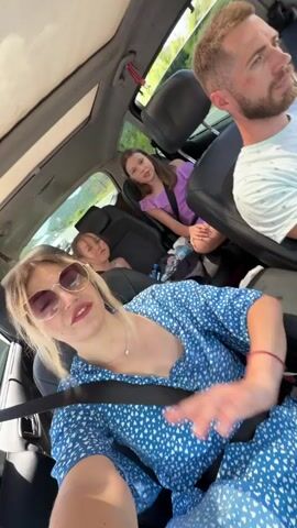 This is a video of a family in a car. The subjects are a woman in the foreground, a man driving, and two children in the back. The woman is wearing a blue dress with white hearts and sunglasses. She is smiling and gesturing with her hands. The man is wearing a white t-shirt and looking to the side. The children are also looking forward.

The scene appears to be a car journey on a sunny day, likely during daylight hours. The setting is inside a car, with the sunroof open, revealing a glimpse of green trees and possibly a mountain landscape outside. This suggests the family is traveling through a scenic area.

The woman in the foreground seems to be documenting the trip with a selfie video, as she is facing the camera and appears to be talking or singing along to music. The overall mood is cheerful and lighthearted, suggesting a family outing or vacation. There is no visible text in the video. The location context provided is Vrela, Montenegro.