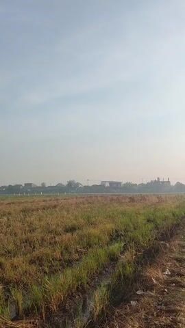 The video shows a road with power lines on the right side, bordered by a field of dry grass on the left. In the distance, there are houses and some greenery. The sky is clear and light, suggesting it is daytime. Two people can be seen walking on the road in the distance. The camera is moving forward, giving the impression of traveling down the road.