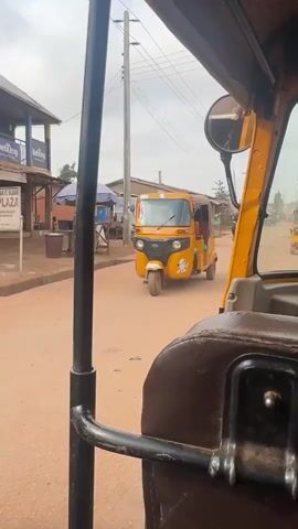The video is filmed from the perspective of someone inside a yellow auto-rickshaw, looking forward. The auto-rickshaw is driving down a dirt road lined with small shops and buildings. Several other auto-rickshaws and a motorcycle are visible on the road. People can be seen walking and standing around the shops. The sky is overcast. The video appears to be taken in a busy marketplace or town.