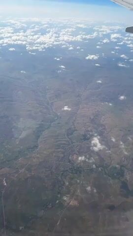 An aerial view from an airplane window shows a vast, open landscape below. The terrain is characterized by rolling hills and a winding river or valley cutting through the land. Patches of green vegetation are interspersed with drier, brown earth, suggesting a semi-arid or rural environment. Scattered white clouds drift across the sky, some casting shadows on the ground. The horizon is visible, with a clear blue sky above and the wing of the airplane extending into the upper right corner of the frame. There are no people or discernible man-made structures visible in detail, though faint lines resembling roads or pathways can be seen on the ground. The overall impression is one of a scenic, natural landscape viewed from a high altitude.