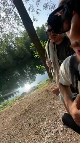The video shows two young men standing next to a body of water, possibly a river or lake. The man in the foreground is wearing sunglasses and a white t-shirt and is holding a cigarette or vape pen. He is looking at the camera and sticking his tongue out slightly. The second man is standing behind him, also wearing sunglasses and a towel draped over his shoulders. He is looking towards the water. The background is a natural scene with trees and sunlight reflecting on the water. The ground in front of the men appears to be dirt and gravel.