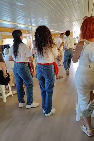 A group of people are standing in a room with a light-colored ceiling and wood-patterned floor. The room appears to be on a boat, with windows visible on the left side. Two women in the foreground are wearing white t-shirts with a red sash tied around their waists and blue jeans. They are facing away from the camera. A woman with red hair is on the right side of the frame, also facing away from the camera and wearing white clothing. In the middle ground, several men and women are standing and appear to be dancing or moving to music. Some are wearing white t-shirts and shorts, while one man is wearing a red t-shirt. The overall atmosphere seems to be festive.