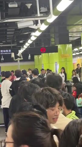 The video shows a large crowd of people lined up at what appears to be an entrance gate, potentially for a subway or train station. A sign above the entrance gate reads "Entrance Gate" in English and Chinese. The area is well-lit with overhead fluorescent lights and has a modern, clean design. The people in the crowd are diverse in age and attire, with many wearing backpacks or carrying bags. There is a security camera visible above the entrance gate, and a red circular sign is illuminated nearby. People are standing closely together, indicating a crowded environment, and some are looking forward toward the entrance. The video appears to have been taken from within the crowd, looking toward the entrance.