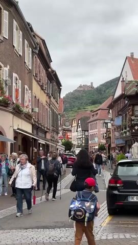 The video shows a street scene with people walking and cars parked. The buildings on either side of the street are old and have unique architecture. In the background, a castle or fortress is visible on a hilltop. The sky is overcast. People are dressed in casual clothing, and some are carrying bags or using walking sticks. The street is paved with cobblestones. The overall atmosphere appears to be that of a tourist town or village.