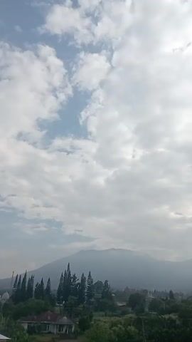 The video shows a landscape with mountains and a cloudy sky. The mountains are covered in vegetation. There are trees in the foreground and buildings visible among the trees. The sky is mostly covered with clouds, with some patches of blue sky and sunlight breaking through. The camera pans slightly upwards.