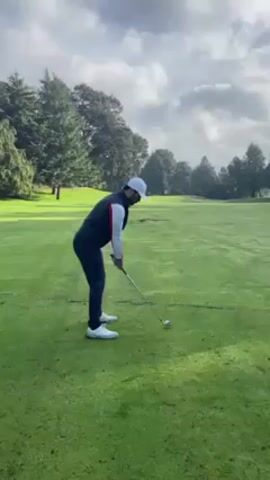 A person wearing a white hat, white and red-striped vest, long-sleeved white shirt, and dark blue pants is playing golf on a green, grassy field. He is seen from the side, bending at the waist with his legs slightly bent. He holds a golf club in his hands and swings at a golf ball in front of him, finishing the swing with the club pointed upwards. The sky is overcast with white and gray clouds, and there are several trees and a patch of dirt visible in the background.