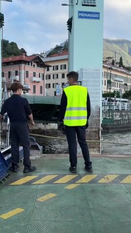 Two men stand on a ferry deck. One man on the left wears a dark navy uniform. The other man on the right wears a neon yellow safety vest over a dark long-sleeved shirt and dark pants. Both men face away from the camera, looking towards the water and a town in the distance. The ferry deck is green with yellow caution stripes. A sign on a white tower above them reads "MENAGGIO." Buildings with pink and white facades are visible on the shore, with hills and mountains in the background under a cloudy sky. The water is choppy. The scene appears to be a ferry terminal.