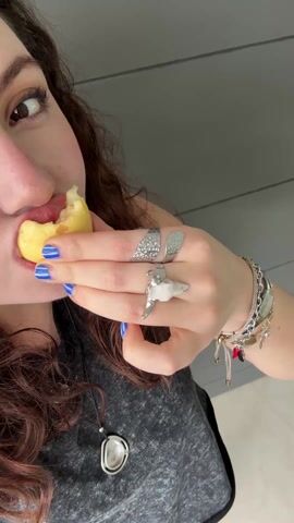A woman with long, curly brown hair, wearing a gray shirt and a pendant necklace, takes a bite from a pale yellow fruit, then holds the bitten fruit close to the camera. She has blue nail polish on her nails and wears several rings and a bracelet. The background is a gray wall.